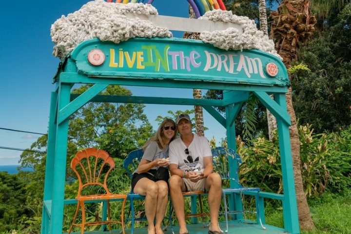 Couple sitting on a colorful bench with 'Live in the Dream' and rainbow sign under palm trees.