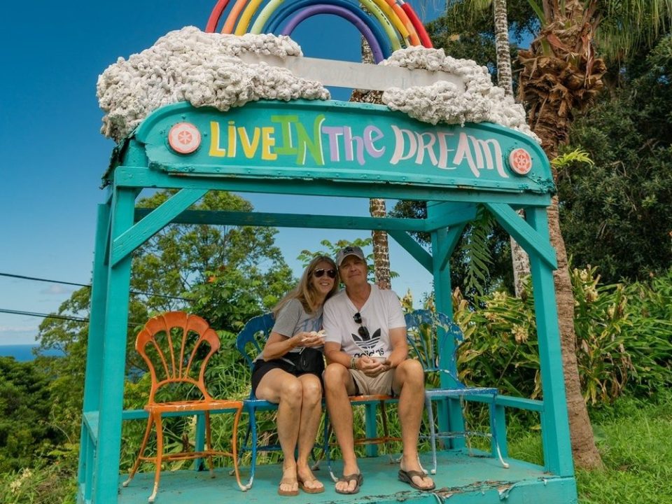 Couple sitting on a colorful bench with 'Live in the Dream' and rainbow sign under palm trees.