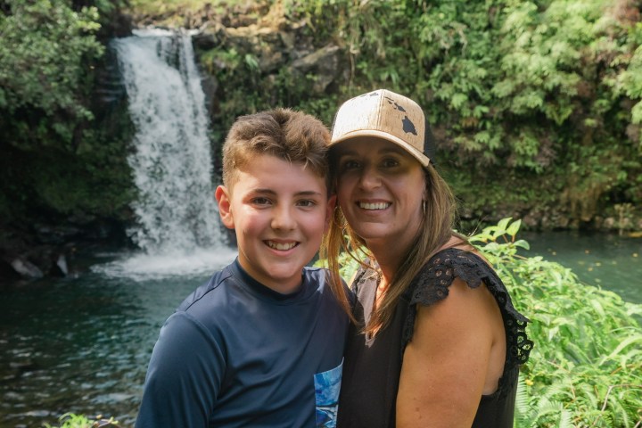 Woman and boy smiling in front of waterfall and greenery.