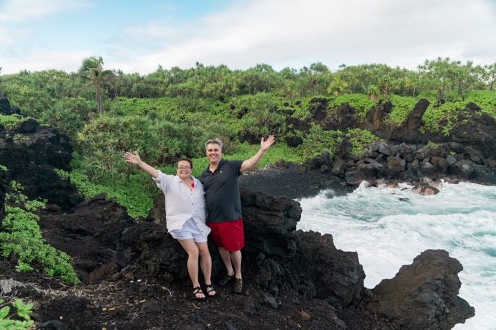 Two people posing on rocky coast with greenery and ocean waves.