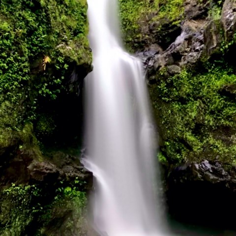 Tall waterfall flowing over lush green rocks into a pool.