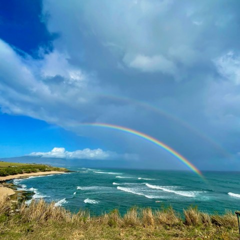 Coastal view with double rainbow over ocean waves under cloudy sky.