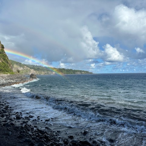 Coastal view with waves, rocky shore, cliff, and a rainbow under a cloudy sky.