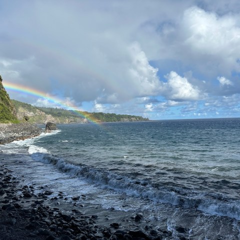 Rainbow over rocky shoreline with ocean waves and cloudy sky.