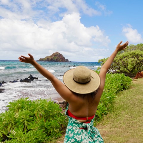 Woman in hat with arms raised, overlooking ocean and rocky landscape.