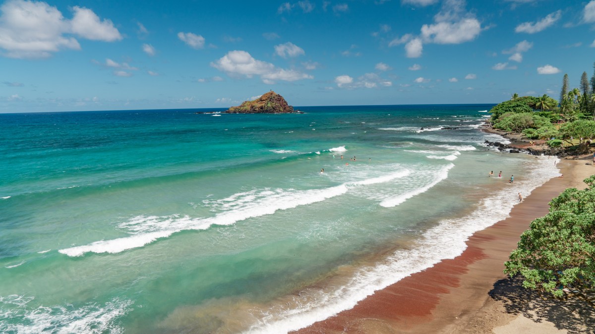 Tropical beach with red sand, turquoise water, and small island in the distance under a blue sky with clouds.