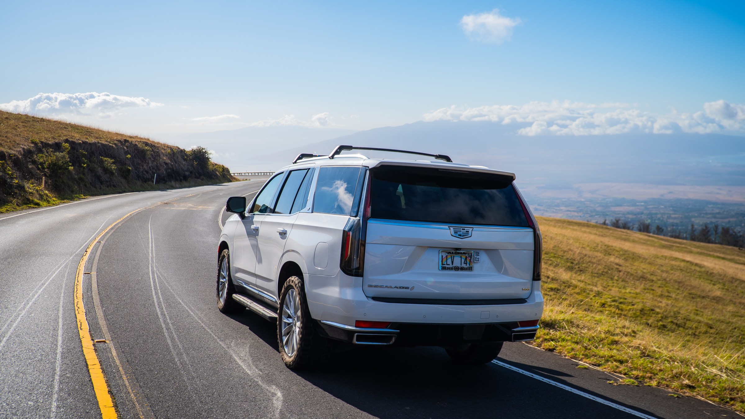 White SUV driving on a scenic, winding road with grassy hills.