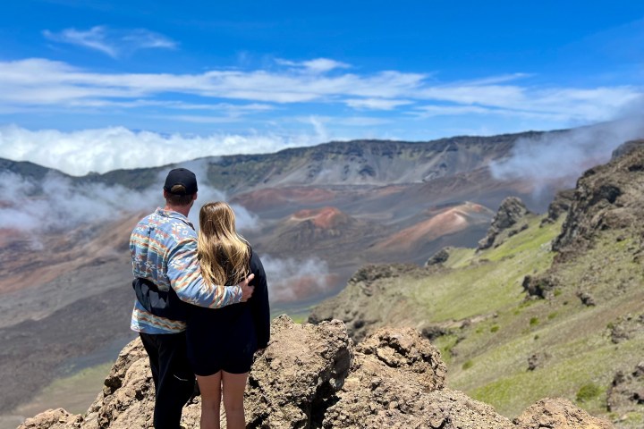 Two people stand on a rocky edge overlooking a vast, mountainous landscape.