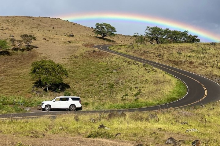 White SUV on winding road under a rainbow in a hilly landscape.