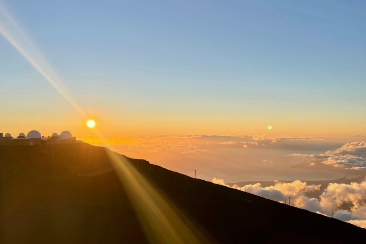 Sunset over mountain with observatories, clouds below.