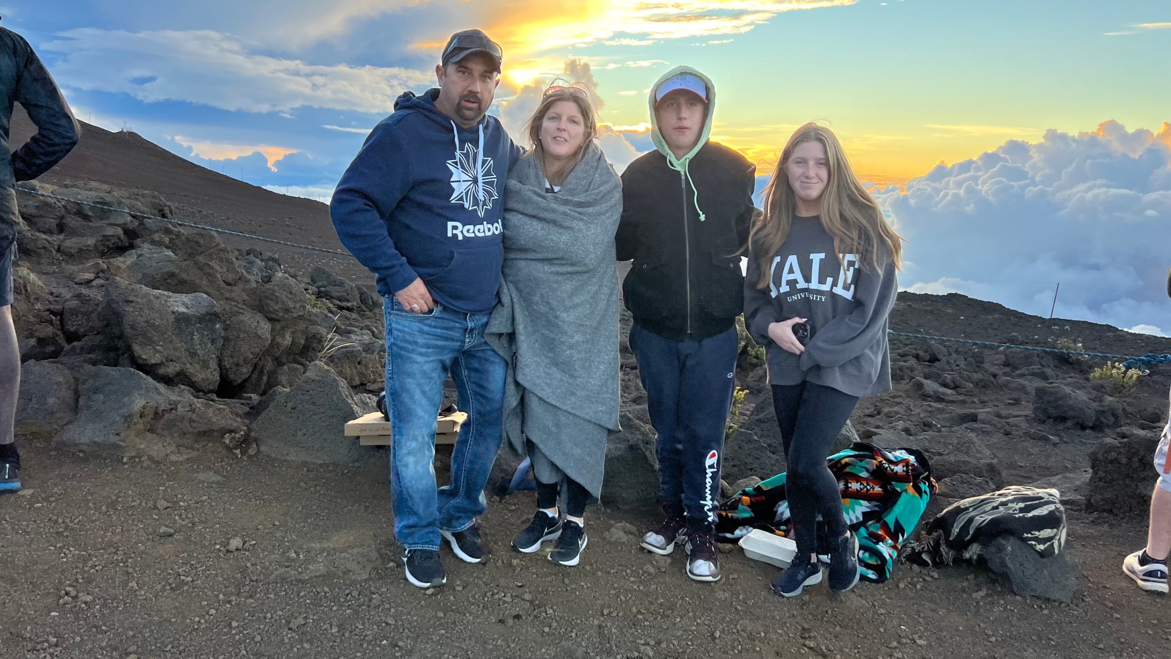 Four people stand on a rocky hilltop at sunset, wrapped in warm clothing.