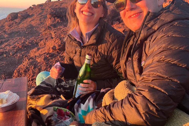 Two people in sunglasses sit on rocky terrain at sunset, wrapped in blankets.