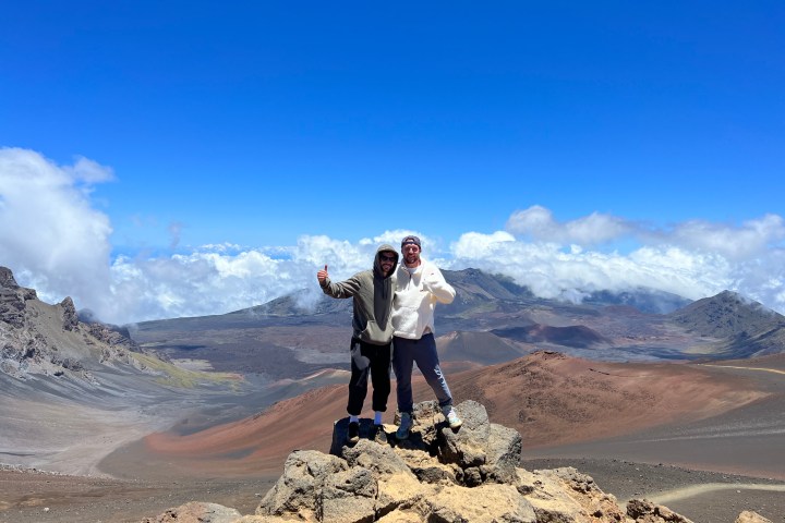 Two people pose on a rocky summit with a mountainous landscape in the background under a blue sky.