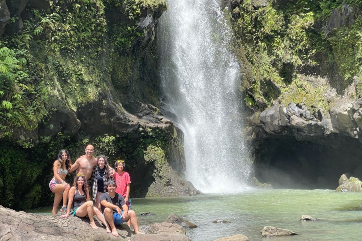 Group of six people posing by a waterfall in a lush, rocky environment.