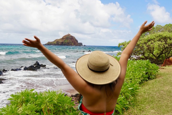 Person in hat and green dress raises arms by a beach with blue sky.