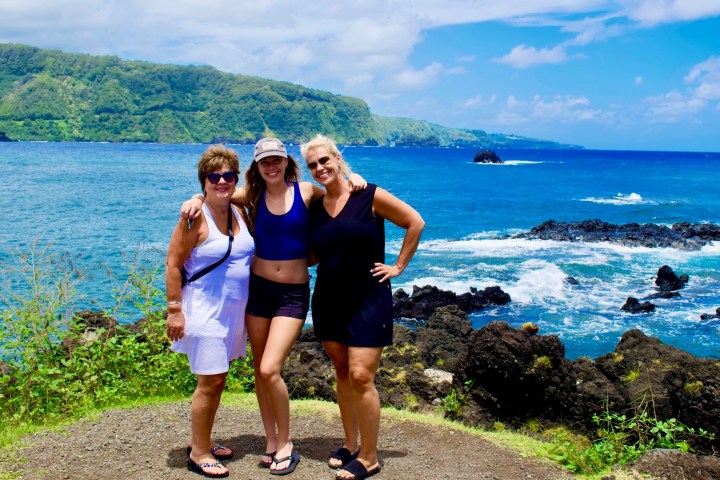 Three women posing by the ocean with lush greenery and blue sky.