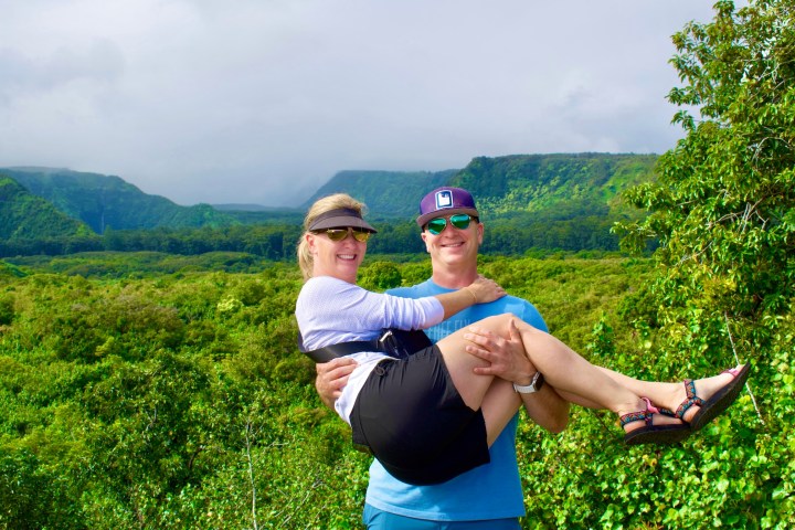 Man carrying woman in front of lush green landscape with hills.