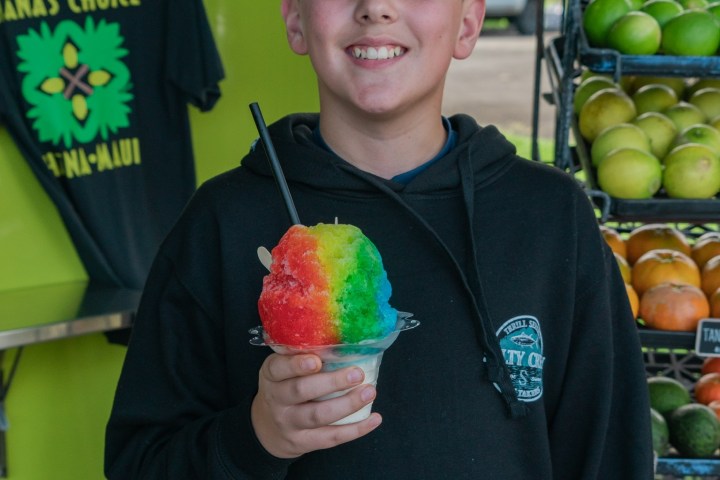 Person holding rainbow shave ice, standing near fruit display.