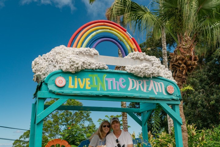Couple sitting under colorful rainbow arch with 'Live in the Dream' sign, surrounded by tropical trees.