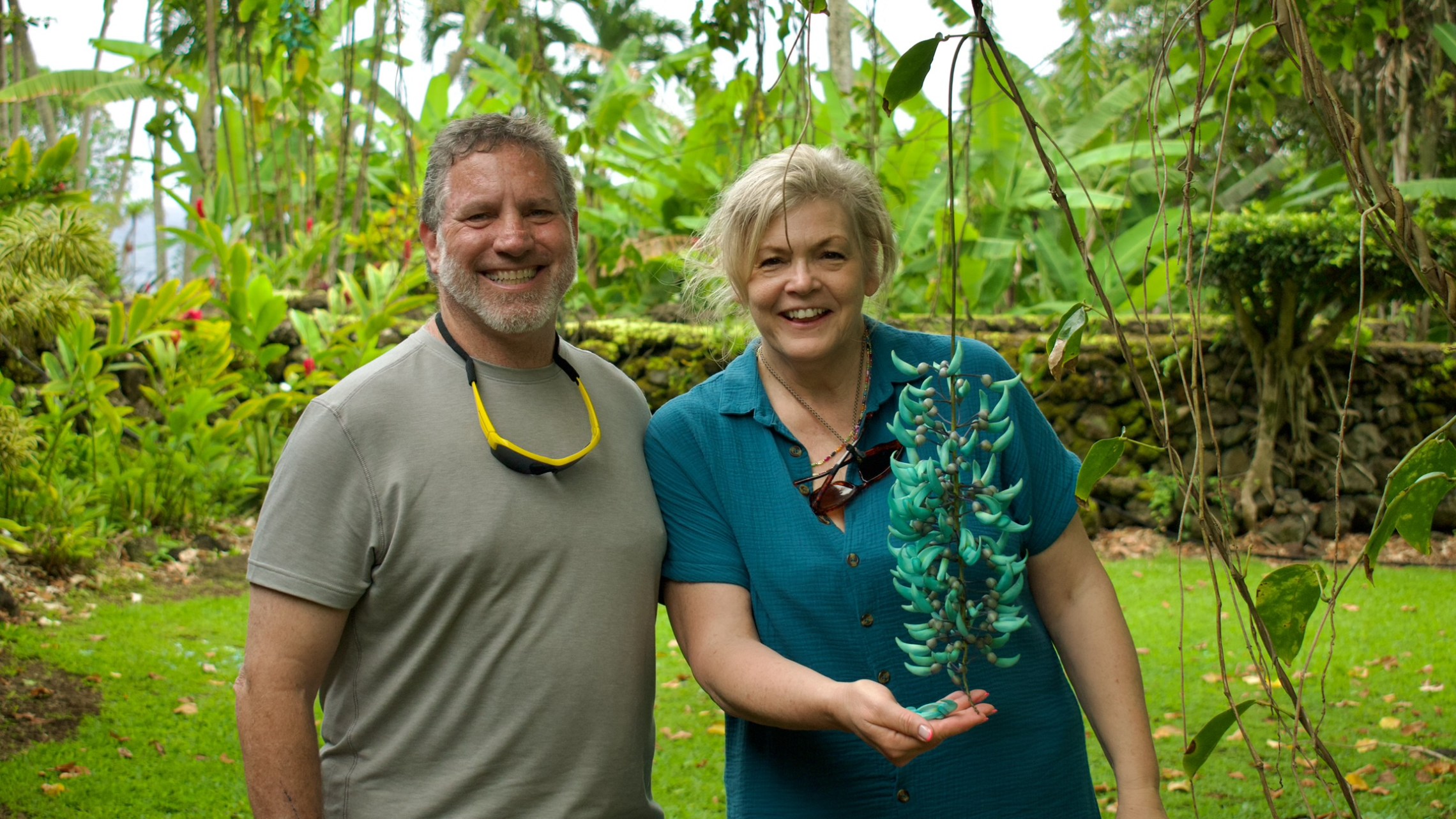 Two people smiling in a lush garden, woman holding a green vine.