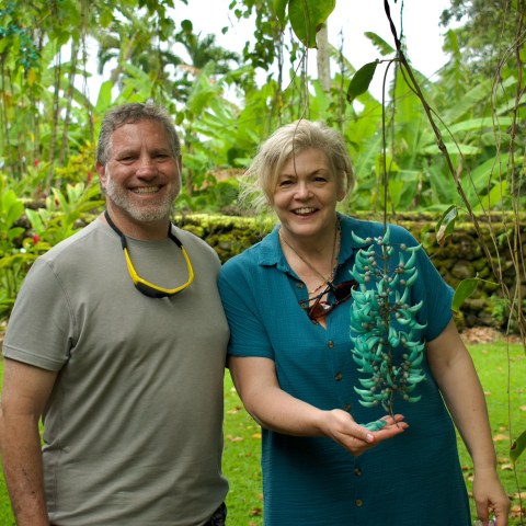 Two people smiling in a lush garden, woman holding a green vine.