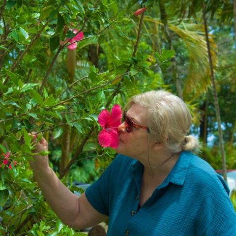 Person with blonde hair smelling a pink hibiscus flower in a garden.