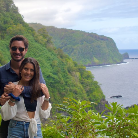Couple smiling in front of lush coastal greenery and ocean view.